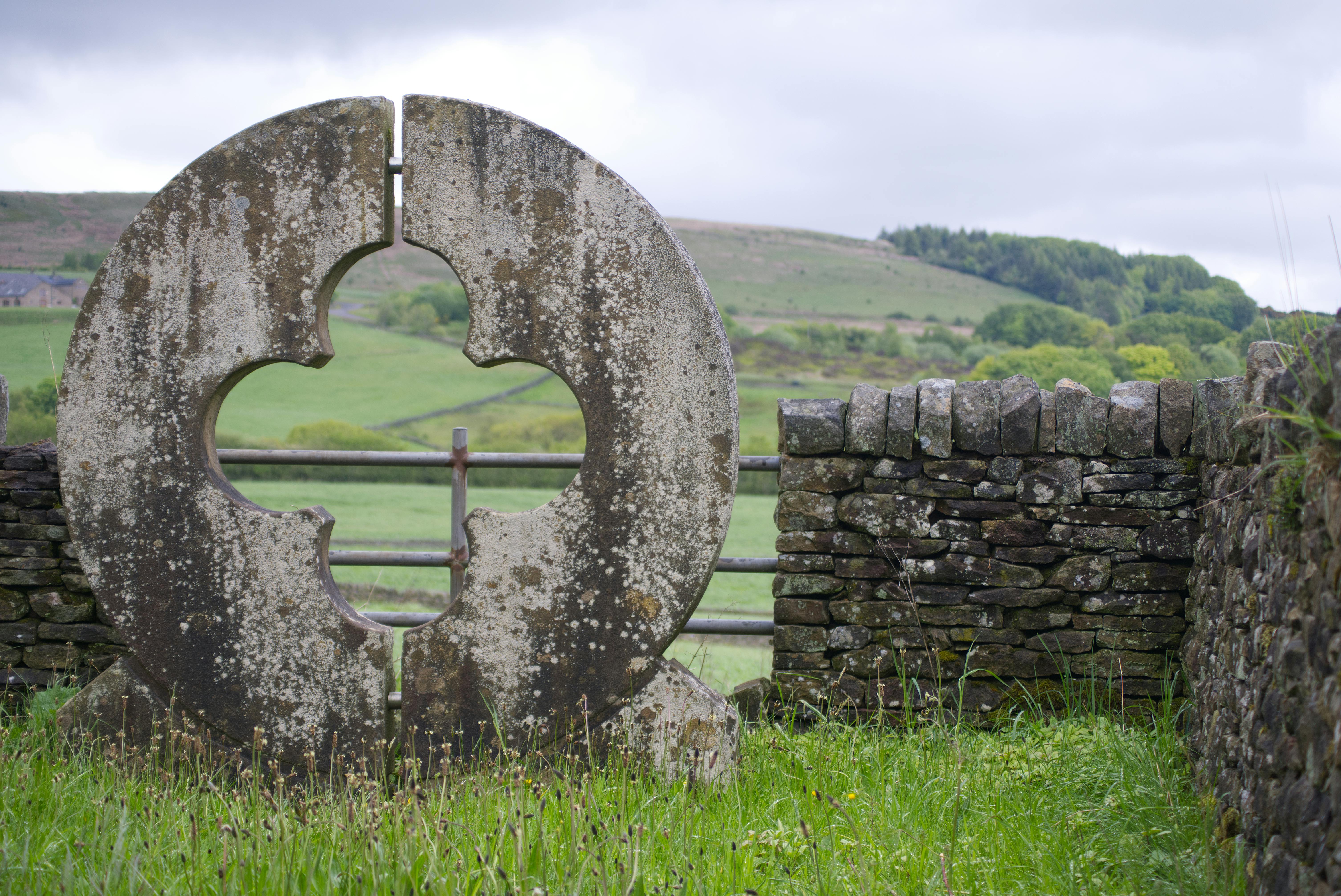 A weathered stone sculpture stands against lush green fields and old stone walls in Horwich, England.