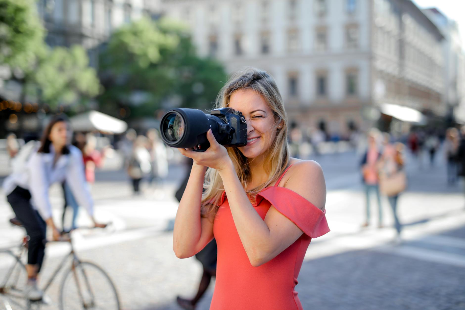 Smiling woman in a pink dress photographs a city street with a DSLR camera, outdoors.