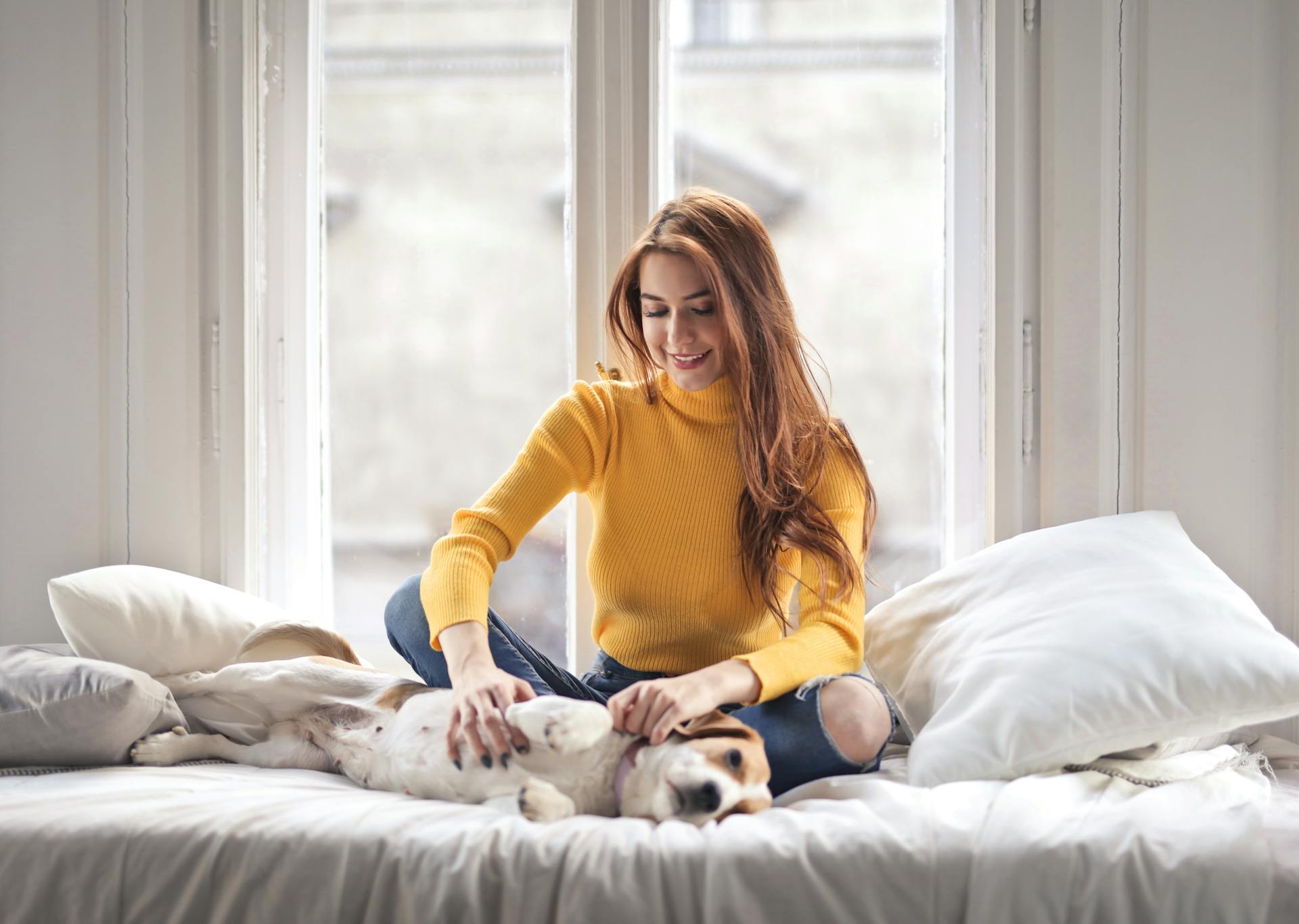 Photo of Smiling Woman in a Yellow Turtleneck Sweater Sitting on Bed Petting Her Dog