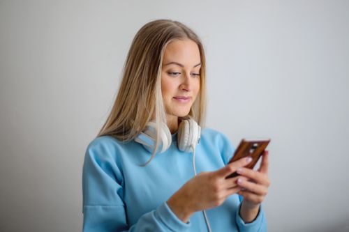 Free Blonde woman in a blue top texting on her phone, wearing white headphones and smiling. Stock Photo