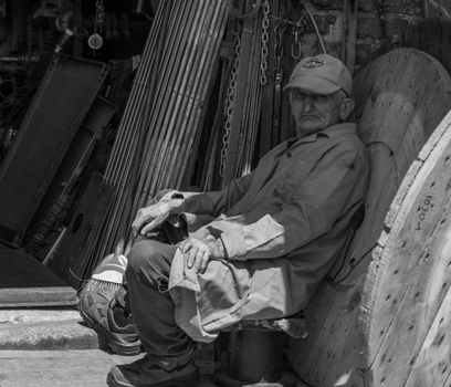 Black and white portrait of an elderly worker sitting outside a workshop surrounded by tools.