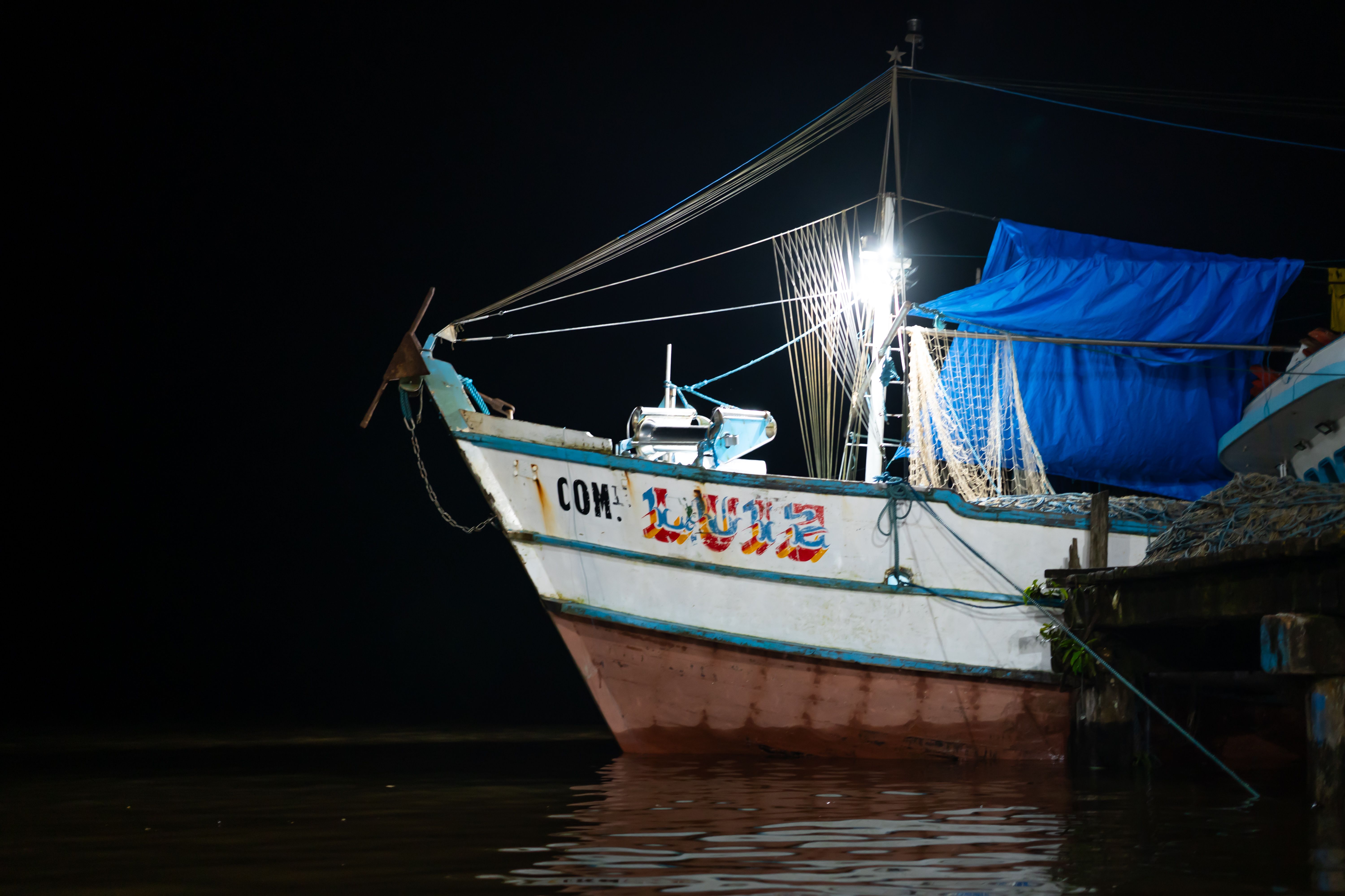 Illuminated fishing boat docked at night, with reflection on calm waters.