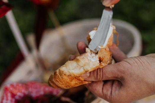 Close-up of hands spreading cream cheese on a croissant during an outdoor picnic.