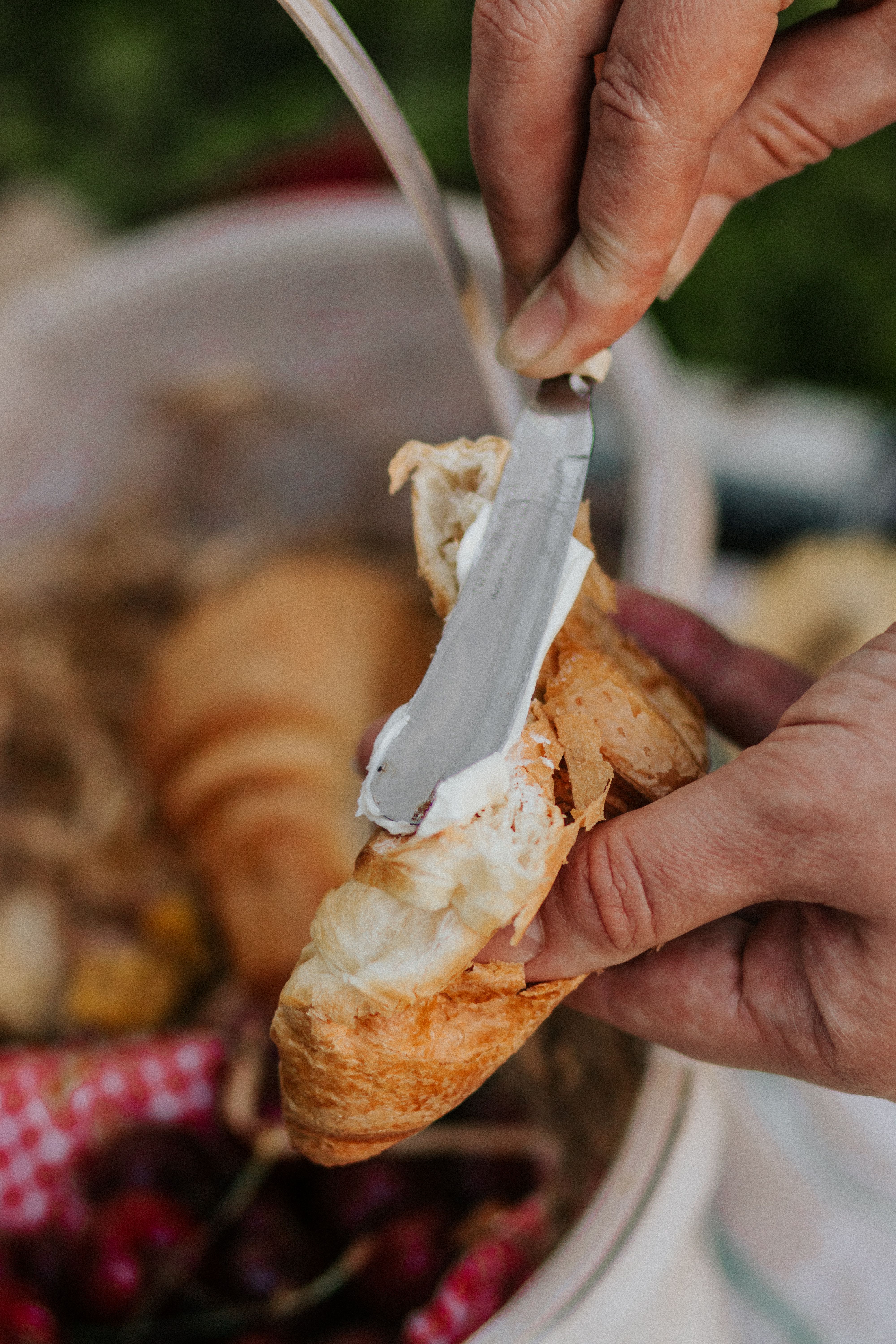A person spreads butter on a croissant with cherries in the background, evoking summer picnic vibes.