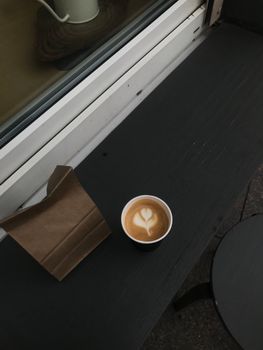A cup of latte with heart art on black table outdoors, near brown paper bag.