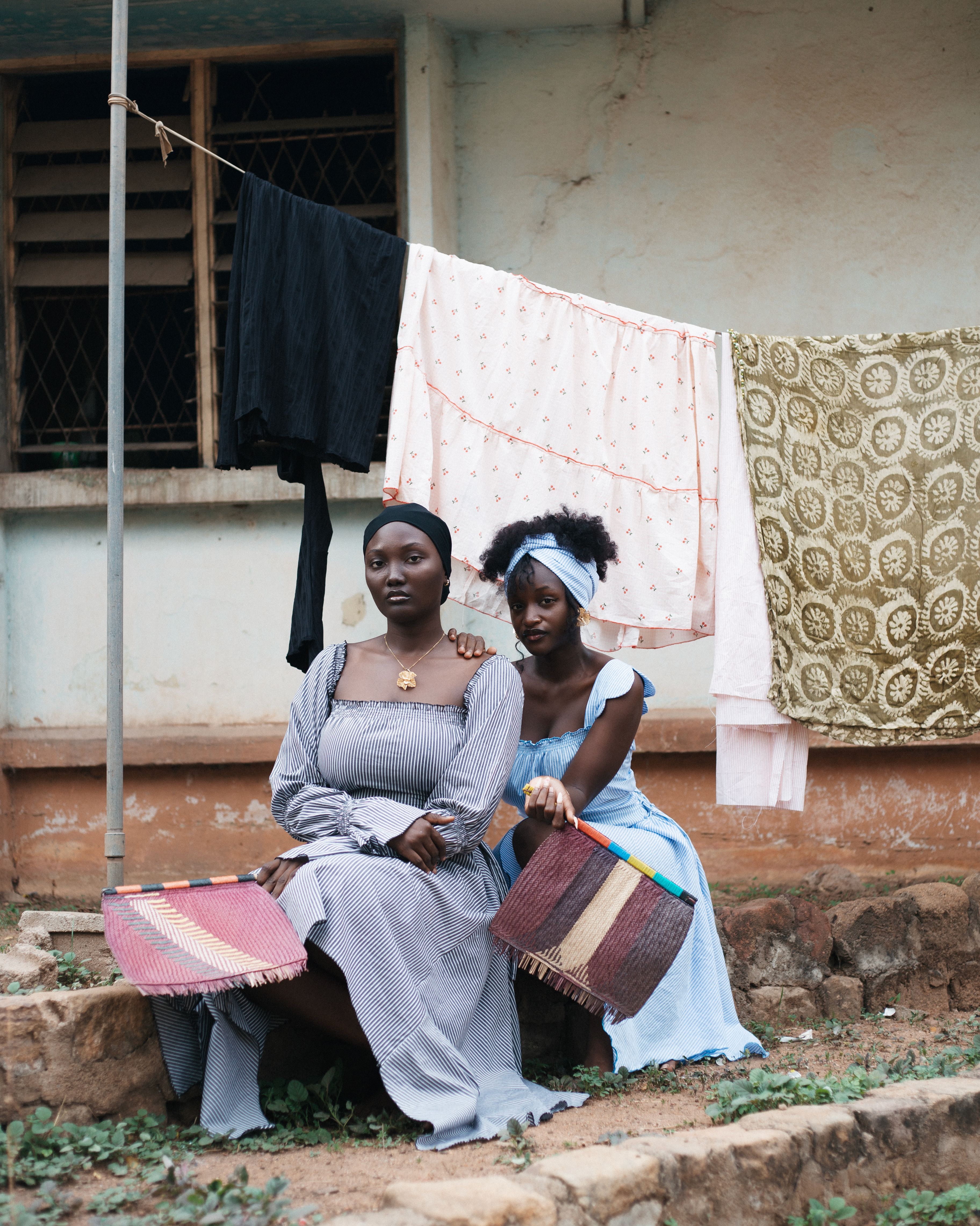 Two Nigerian women sit outdoors in traditional attire with cloths drying in background.