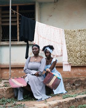 Two Nigerian women sit outdoors in traditional attire with cloths drying in background.