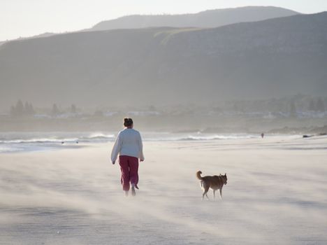 A woman walks her dog along a misty beach in Hermanus, South Africa during a serene day.