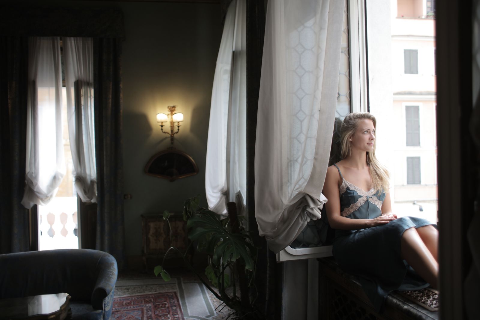 A woman in a blue dress sits by a window, gazing outside, capturing a moment of relaxation.