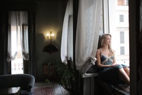 A woman in a blue dress sits by a window, gazing outside, capturing a moment of relaxation.