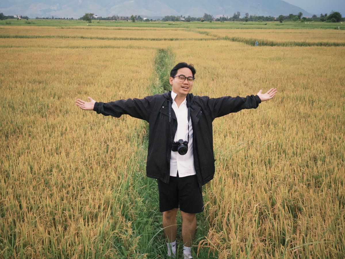 A smiling person standing in a rice field with a camera around their neck, arms open wide