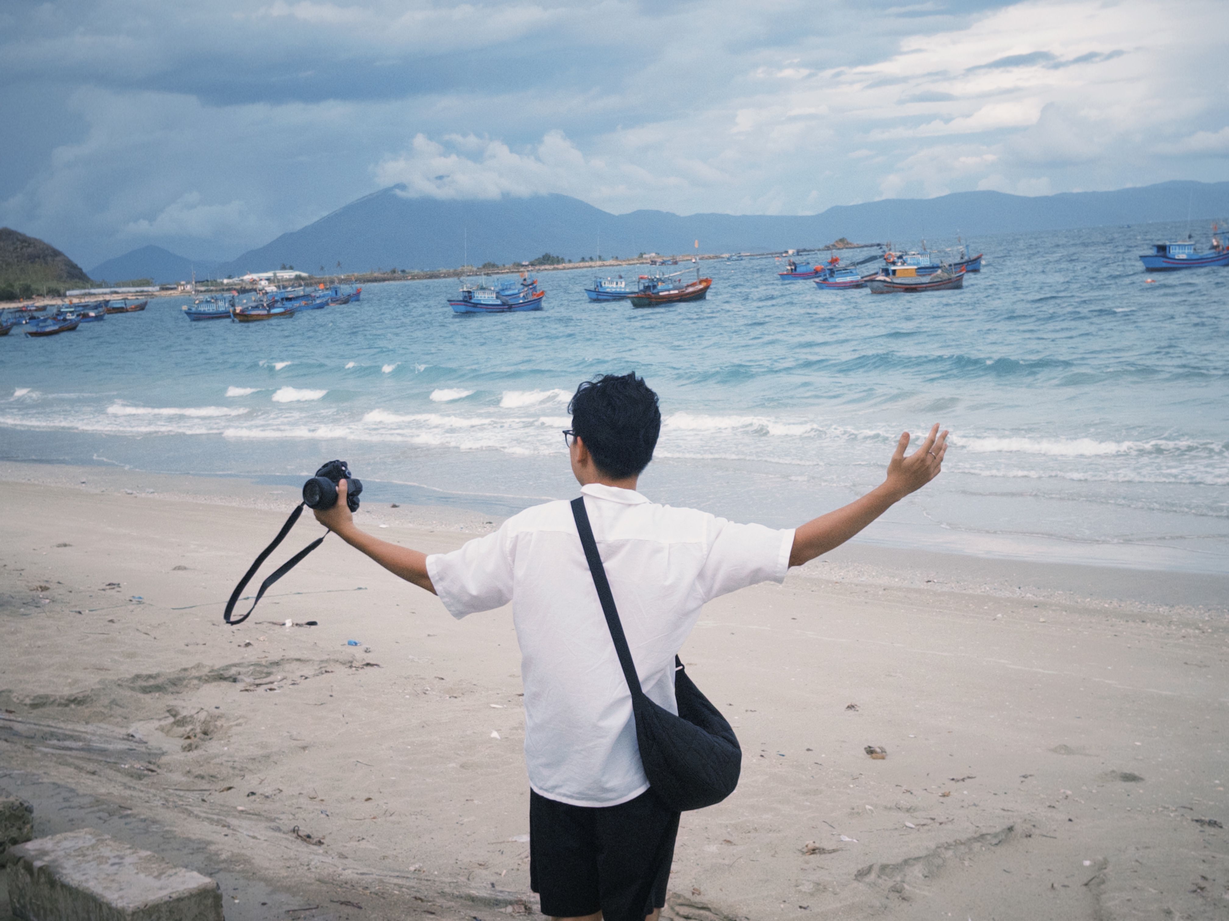 A young photographer stands on a beach embracing the scenic seascape and fishing boats.