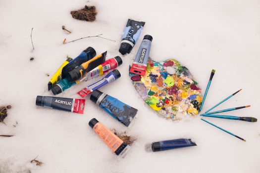 Colorful acrylic paint tubes and palette with brushes lying on a snow surface, artistic winter theme.
