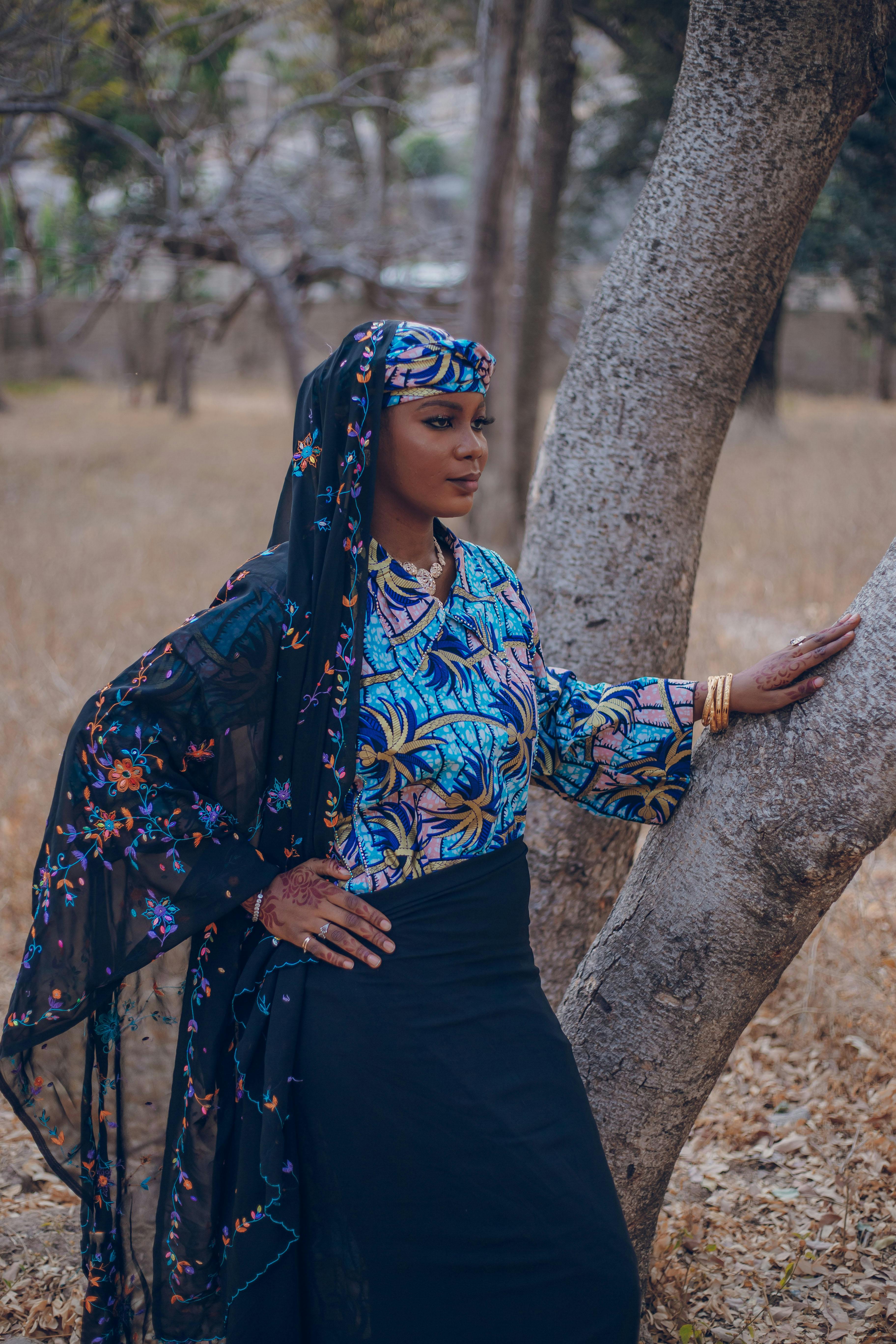 A graceful woman in traditional Nigerian attire stands by a tree within a serene outdoor setting.