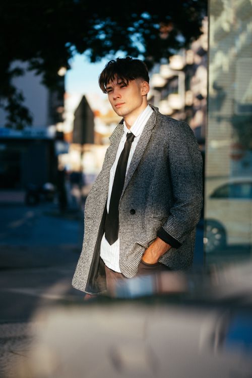 Free Portrait of a young man in smart casual attire leaning against a glass wall outdoors. Stock Photo