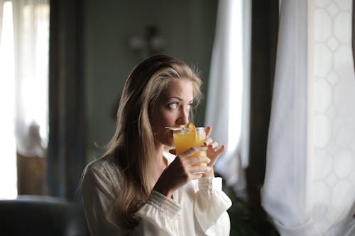 Woman in White Long Sleeve Shirt Drinking an Orange Juice