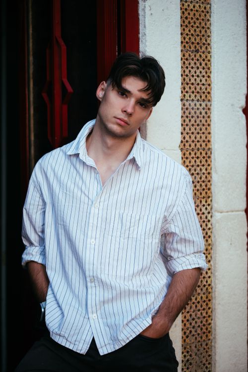 Free A young man in a stylish striped shirt leans against a wall, exuding confidence and modern fashion in an urban setting. Stock Photo