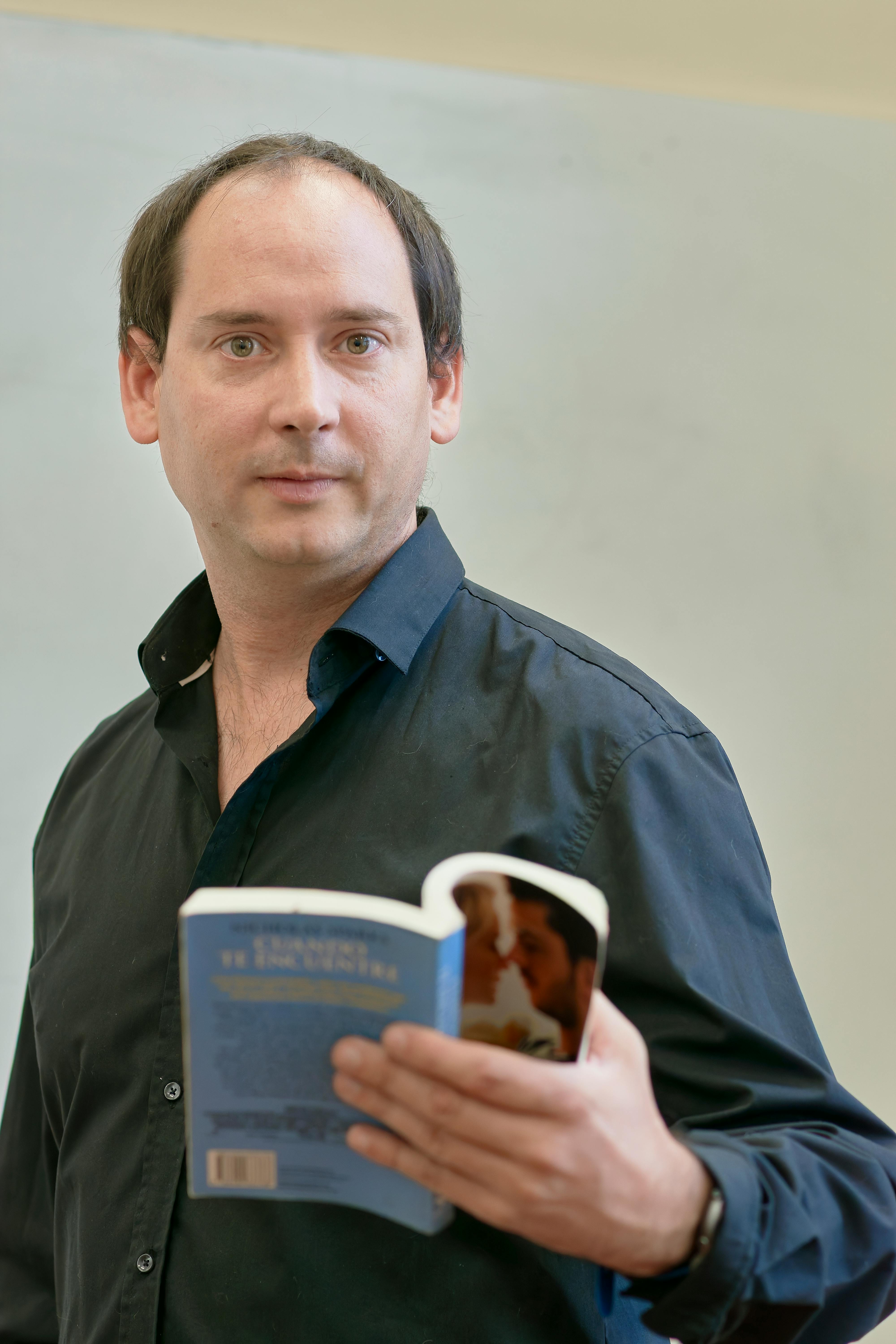 Adult man holding a book with a focused expression, standing indoors.