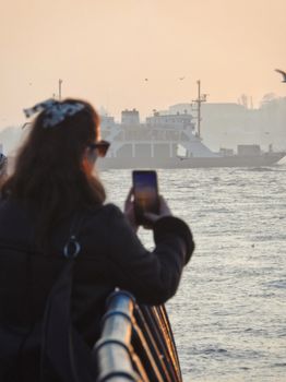 A woman photographs a picturesque river scene with a ferry at sunset, creating a serene and vibrant atmosphere.