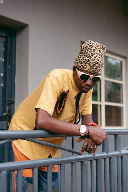 Fashionable adult man with sunglasses and leopard print beanie leaning on a railing outdoors.