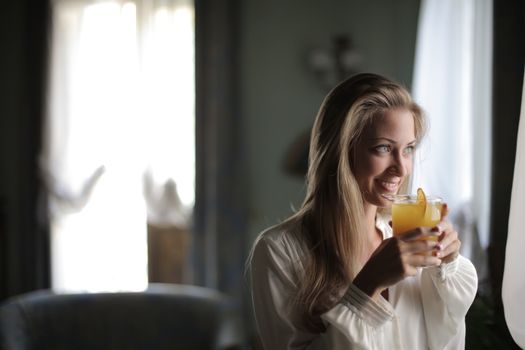 A cheerful woman enjoying a glass of orange juice by a window, capturing a moment of refreshment and relaxation.