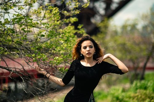 Free A beautiful woman in a black dress poses elegantly outdoors surrounded by lush green foliage. Stock Photo