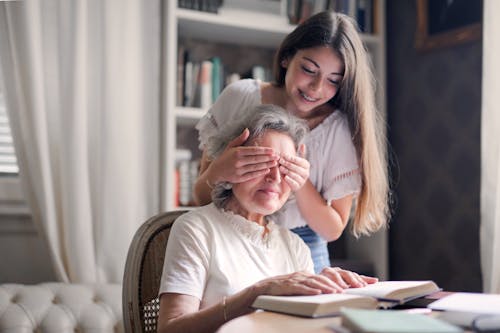 Free From below pensive gray haired woman trying to guess who closing eyes while sitting at table and reading book in light library in cottage Stock Photo