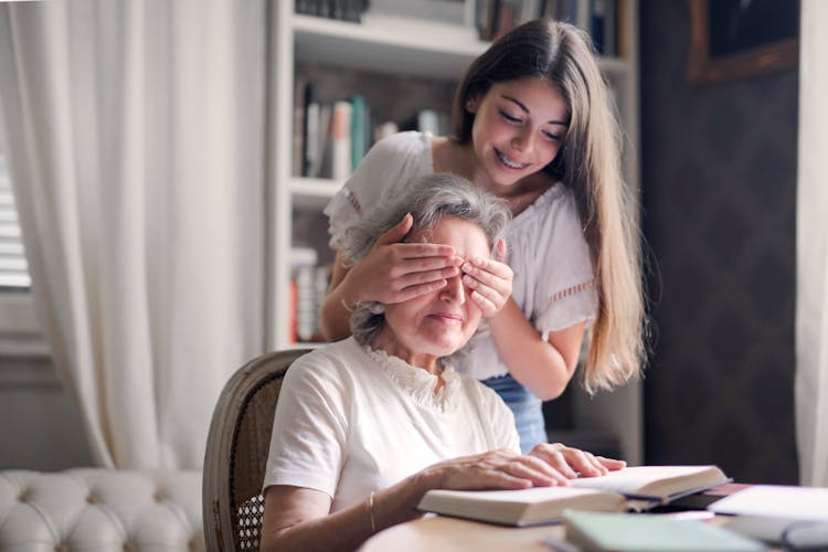 Happy Granddaughter Playing Guess Who Game With Aged Grandmother While Making Surprise In Light Living Room Of Modern Apartment