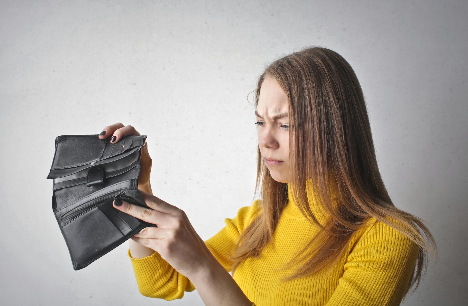 Young woman in a yellow top looking at her empty wallet with a frustrated expression.