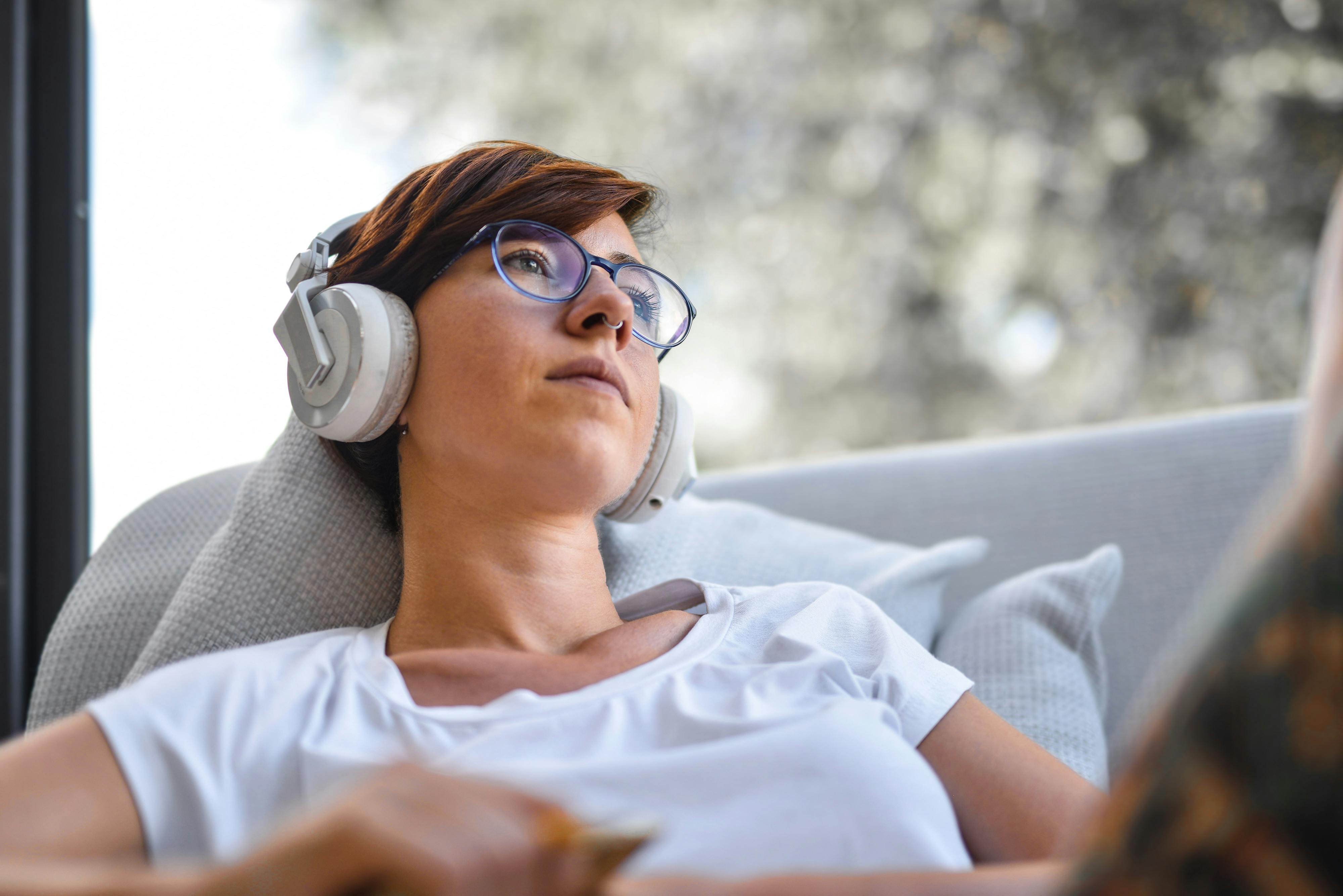 戴眼镜的女人坐在窗前的沙发上，戴着耳机听音乐放松身心