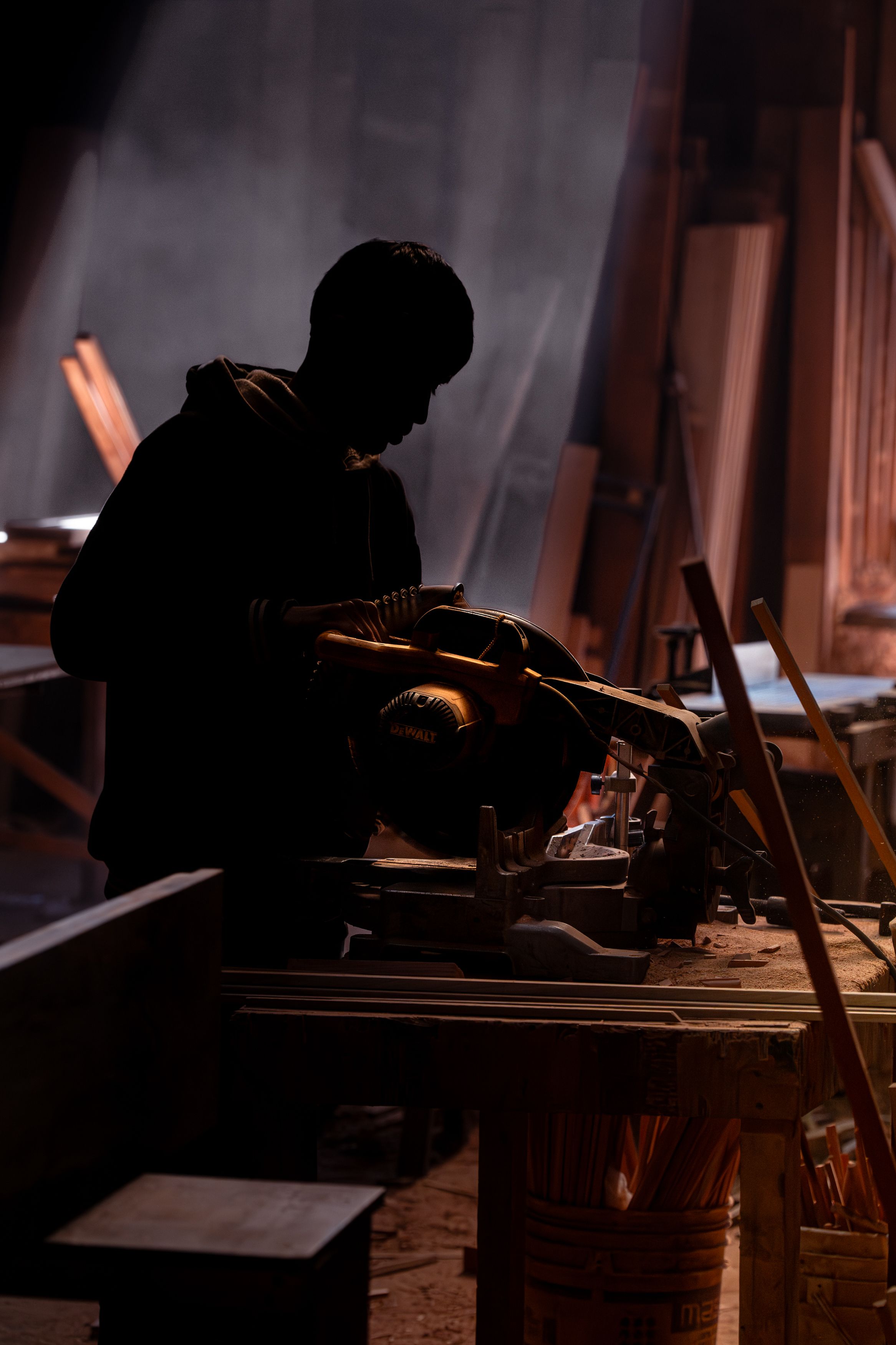Silhouetted carpenter operates a circular saw in a dimly lit workshop, creating a dramatic atmosphere.
