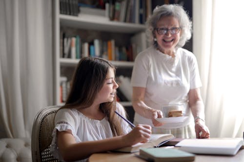 Cheerful grandmother in glasses and casual clothes smiling at camera while giving pastry on plate and mug of tasty beverage to joyful teenager sitting at table with books and exercise book and study in light cozy living room