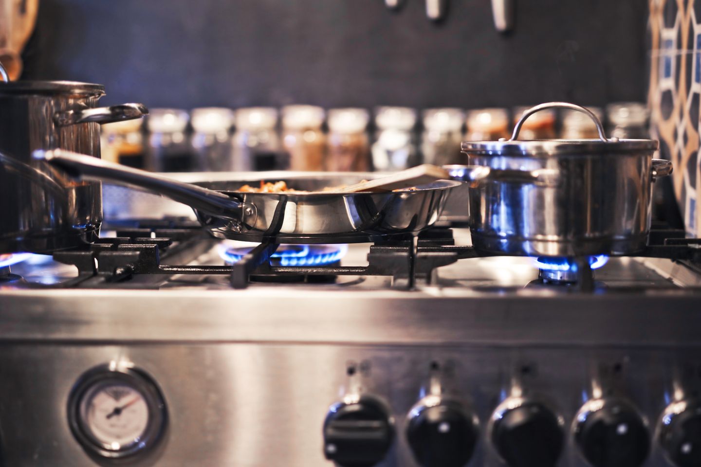 Steel pans over blue gas flames on a working range, with the spice jars lined up behind.