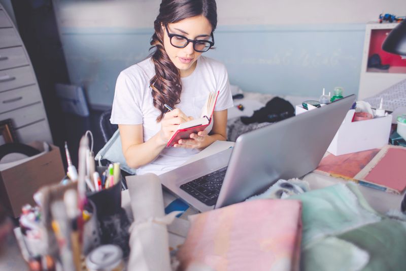 A young woman in glasses studies indoors with a laptop and notepad, writing notes.