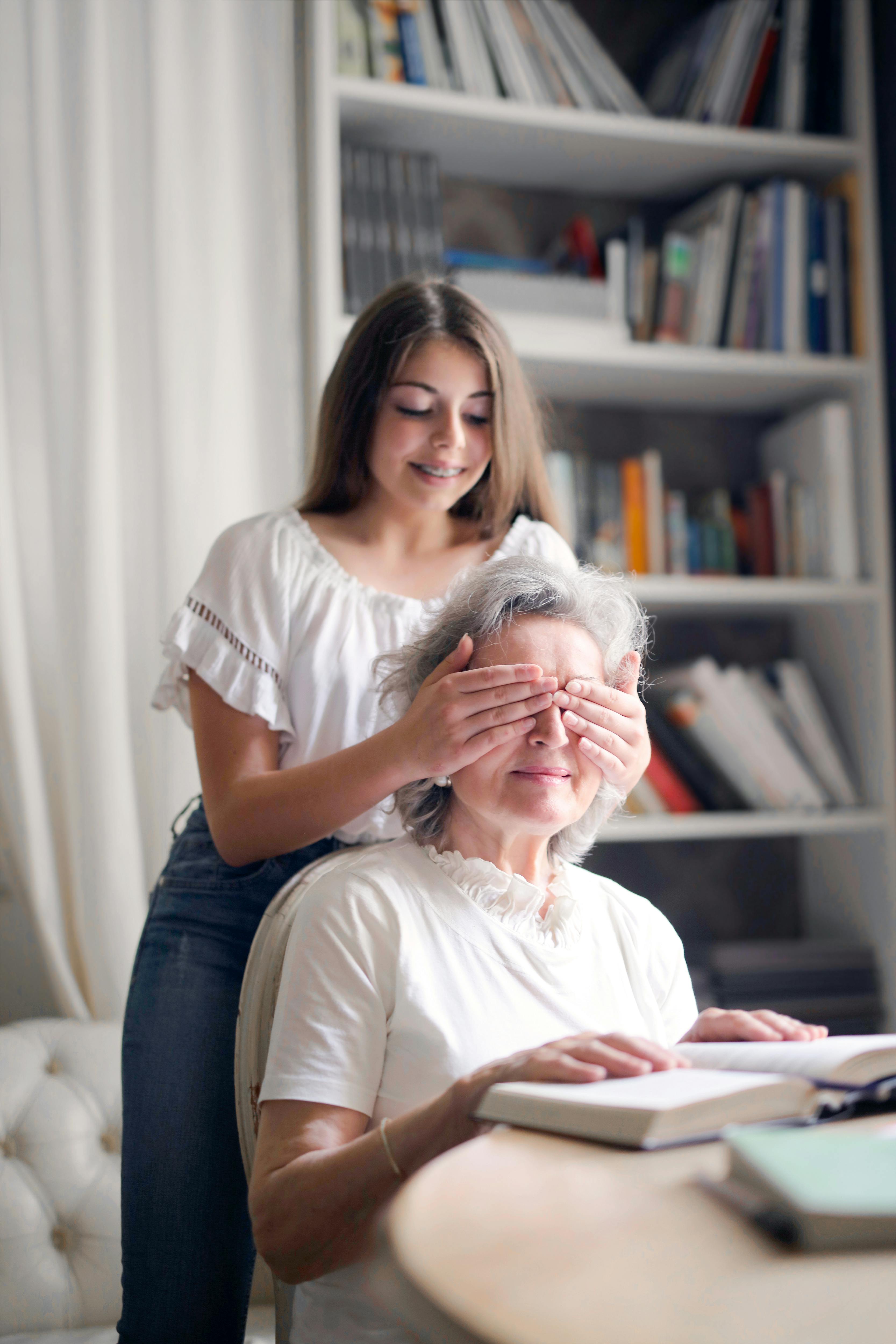 cheerful teenager playing with grandmother guess who game while making surprise in light living room