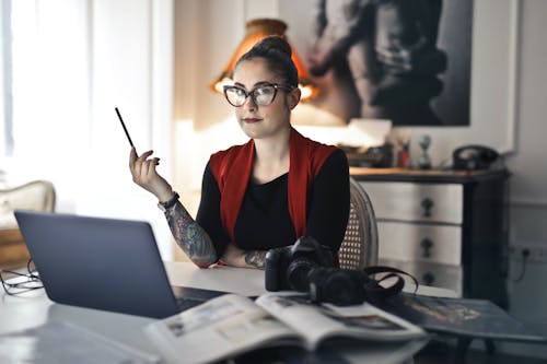Trendy female in glasses and casual clothes looking at camera while sitting at laptop with digital cameras and books against blurred interior of light modern living room