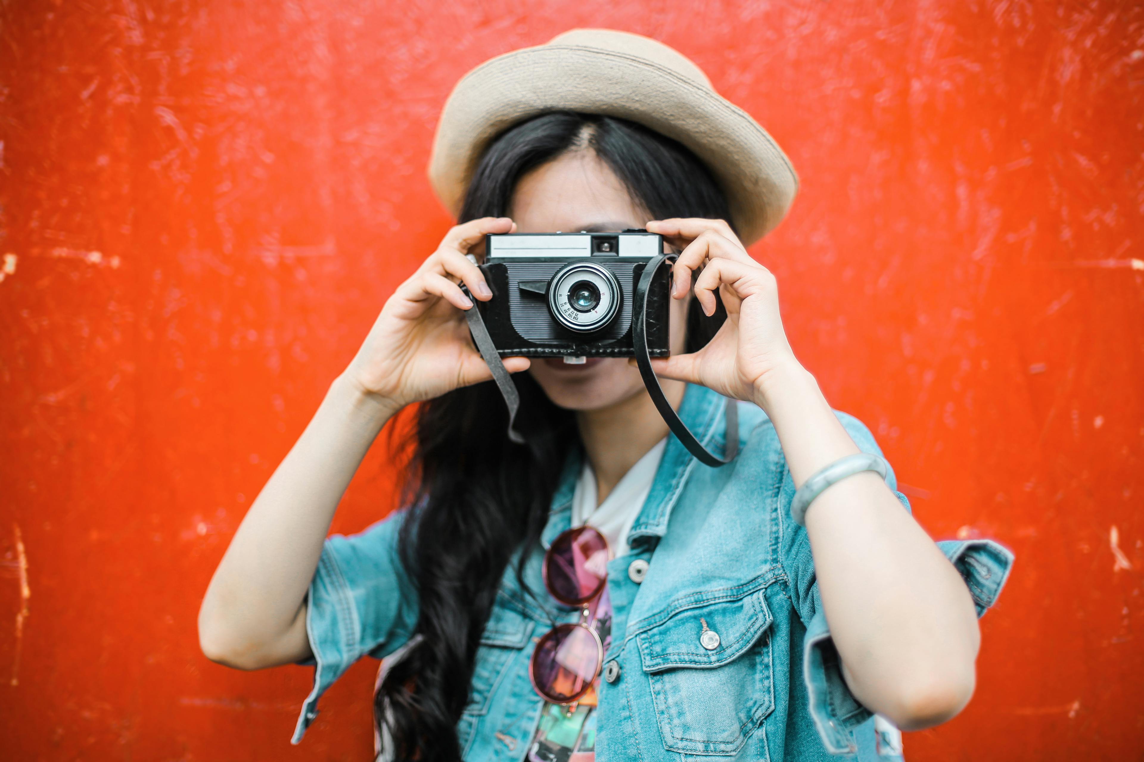Stylish woman in hat and denim holding an analog camera with a vibrant red background.