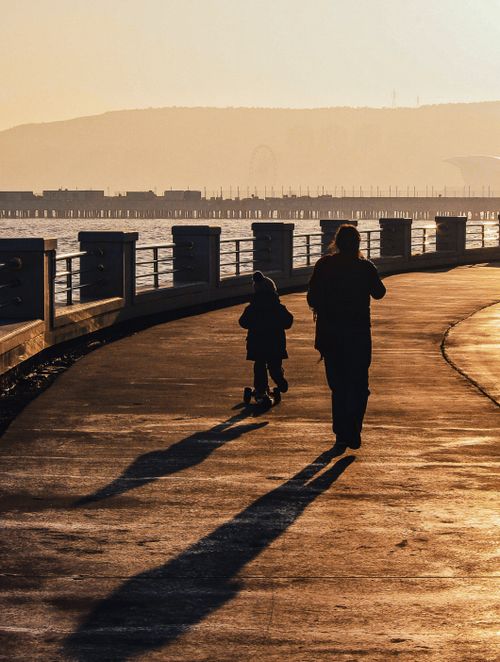 Free Silhouetted figures walking along a waterfront path at sunset with long shadows. Stock Photo