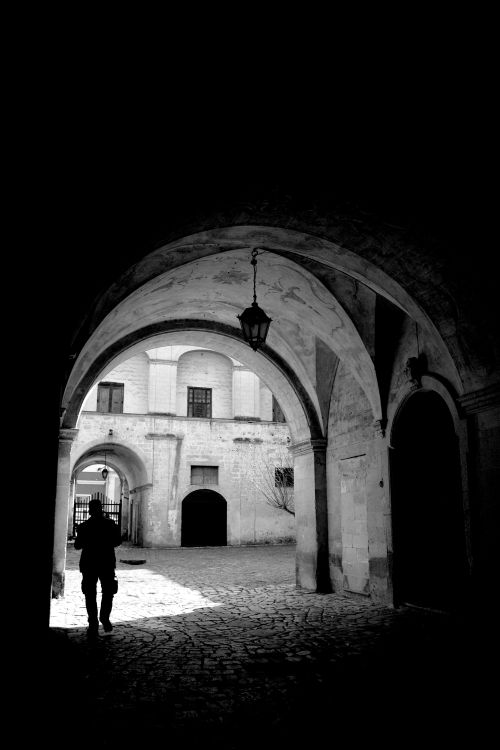 Free Black and white silhouette of a person walking through a historic archway, creating a dramatic urban scene. Stock Photo