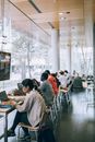 Thoughtful young lady in casual clothes working on laptop while sitting at table near other multiracial visitors against window wall in light modern library