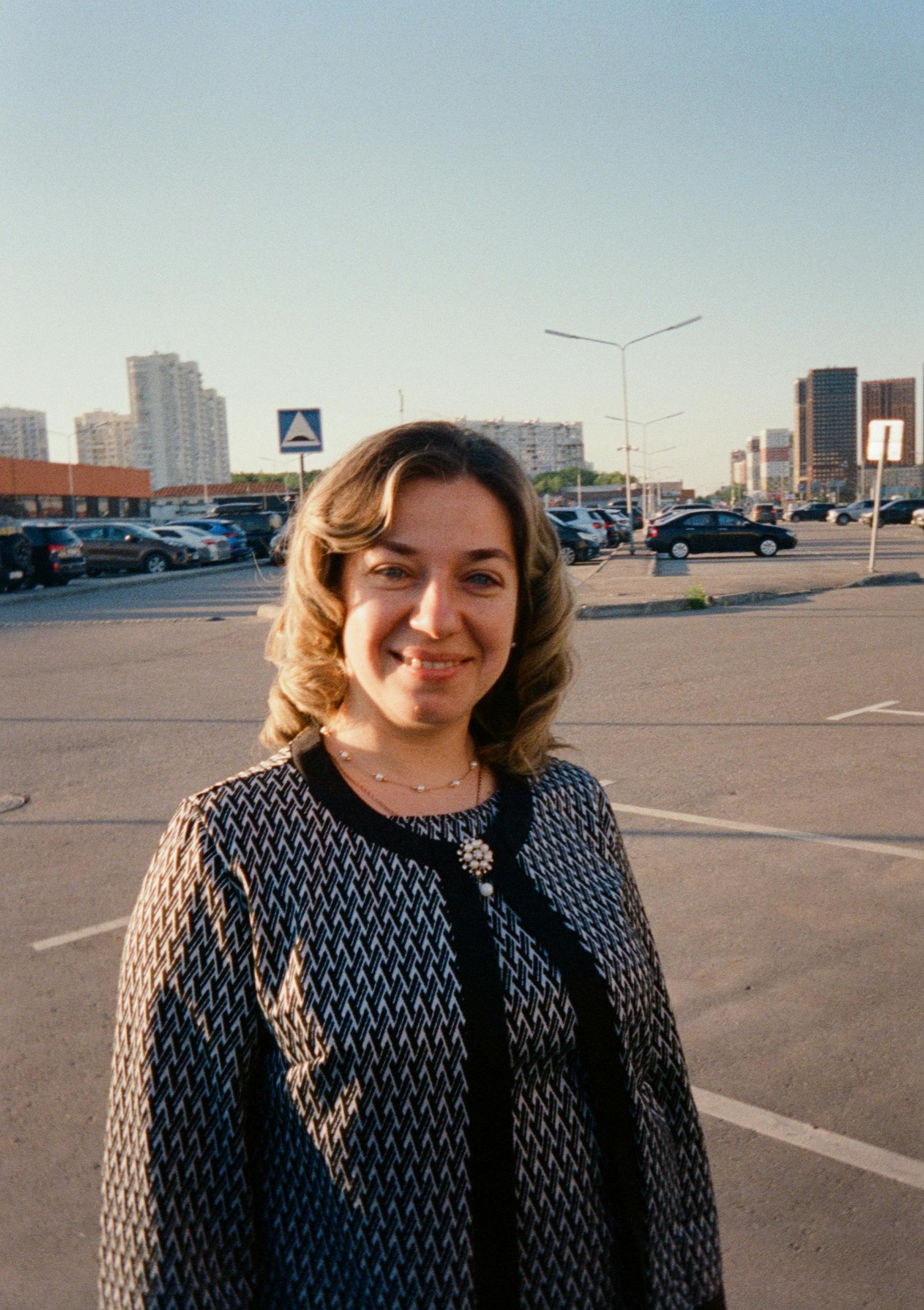 A woman smiling in an urban parking lot with cityscape background, under a clear sky.