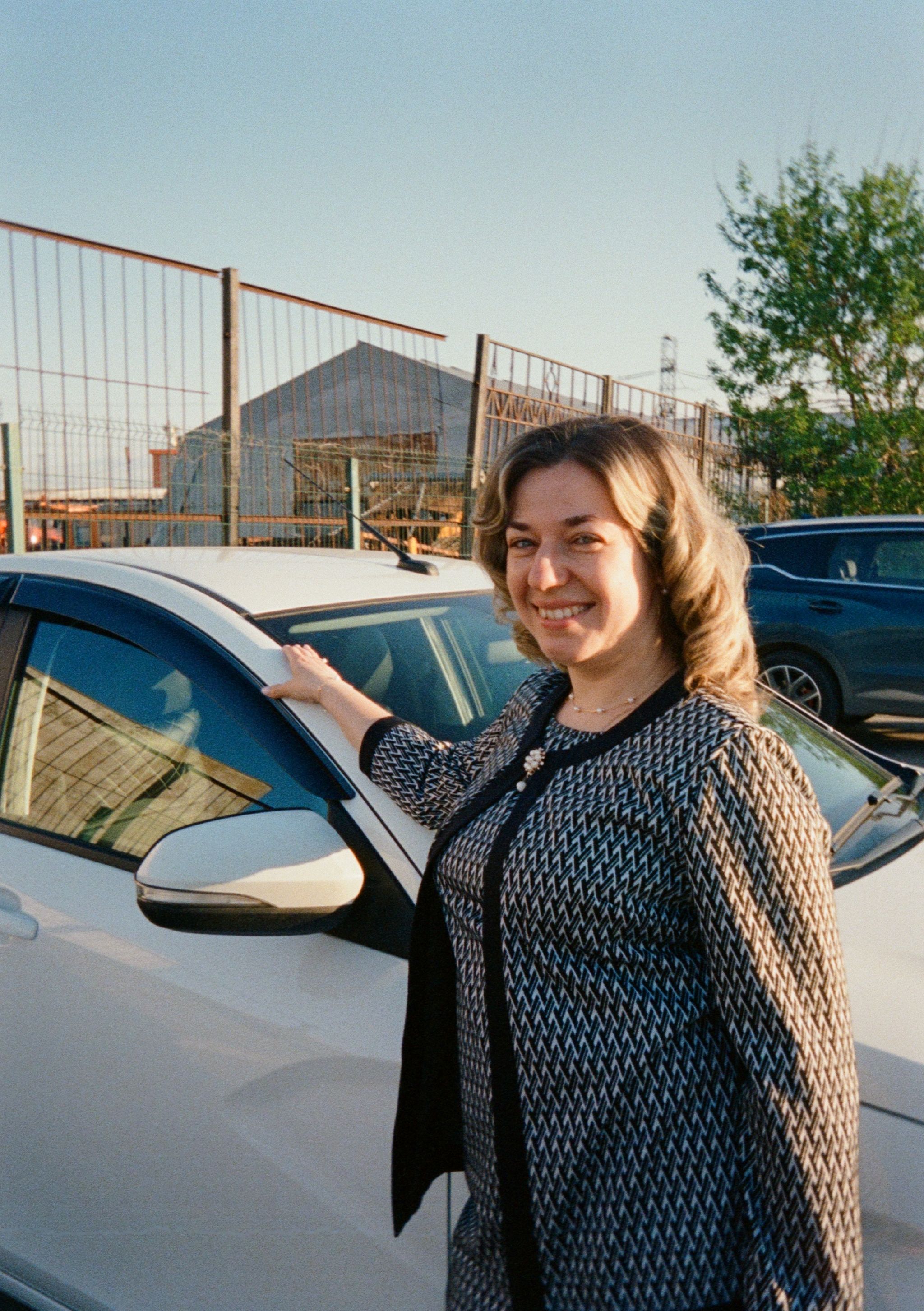 Happy woman in stylish outfit poses by car in sunny outdoor setting.