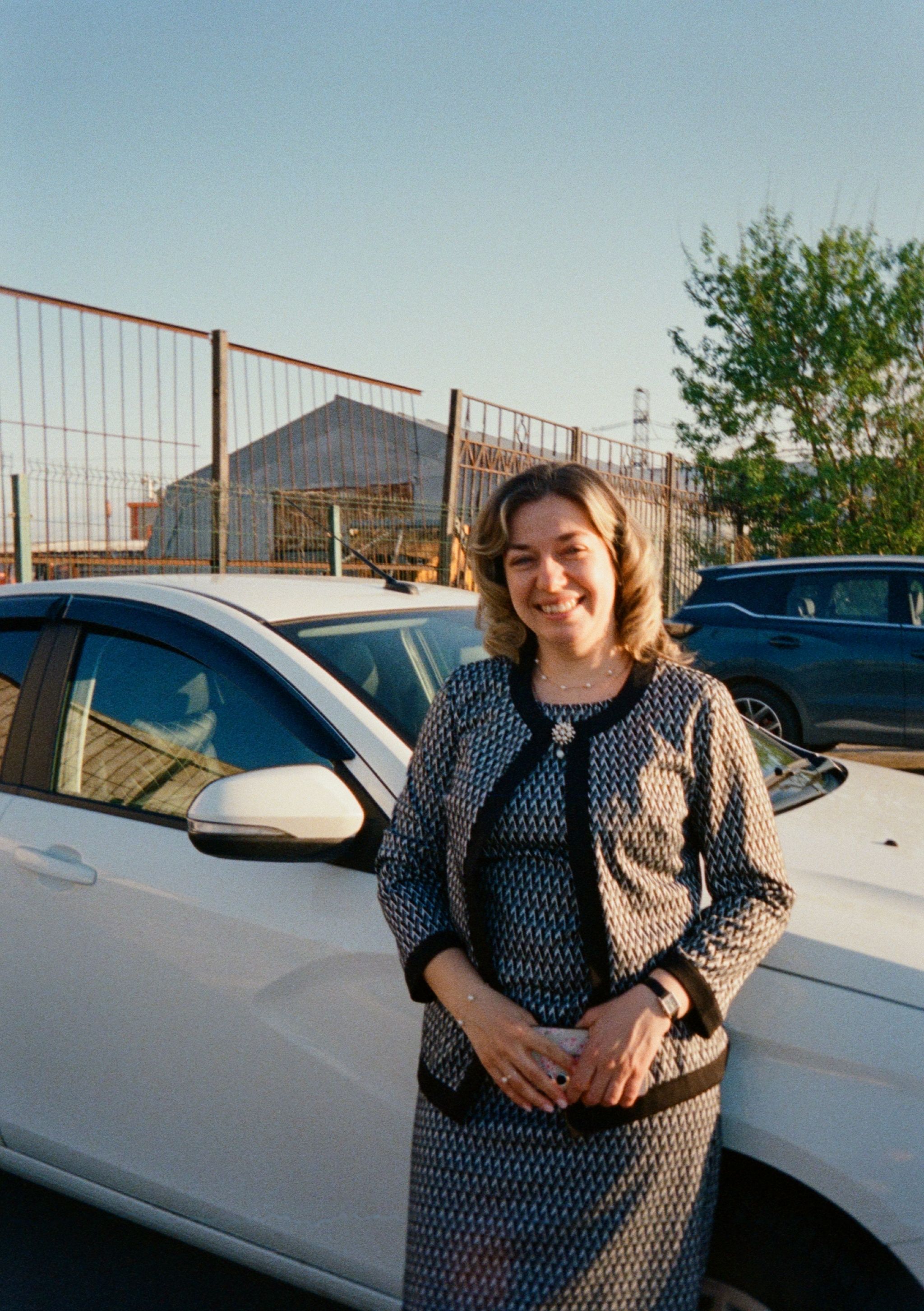A woman dressed in formal attire smiles while standing by a parked car outdoors.