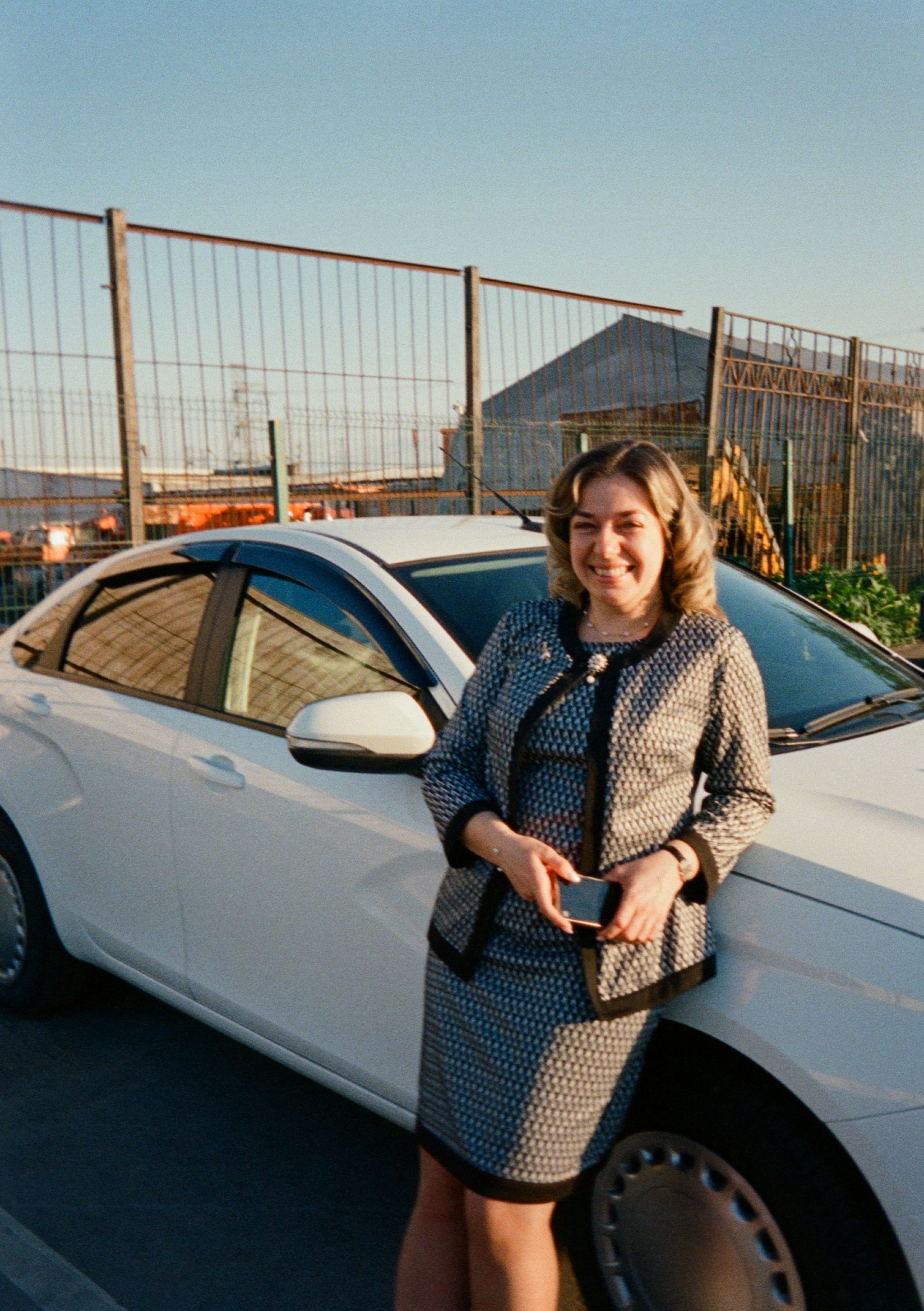 Confident businesswoman in formal attire standing beside a white car outdoors, smiling at sunset.
