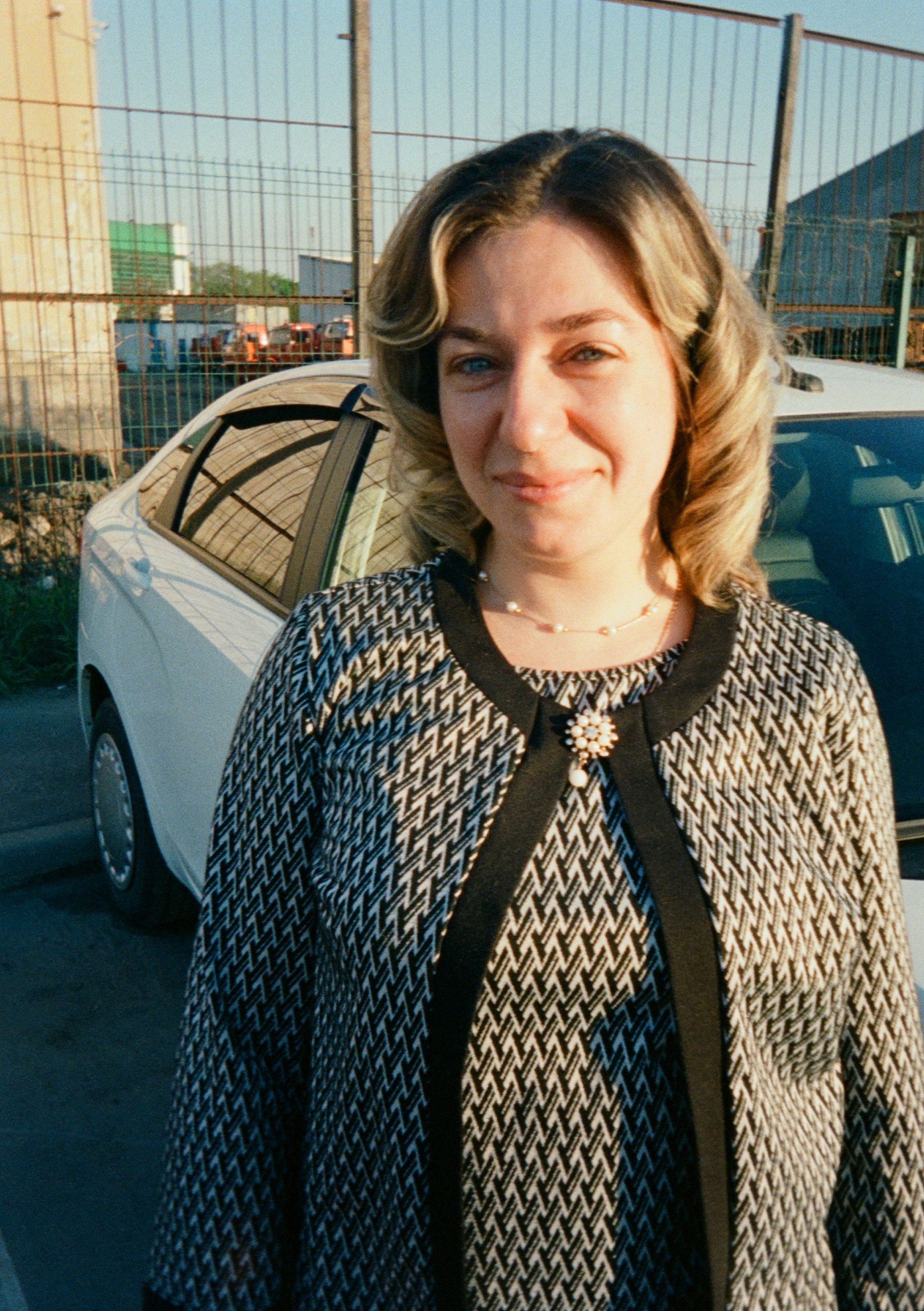Smiling woman standing outdoors with a parked car in the background during daytime.