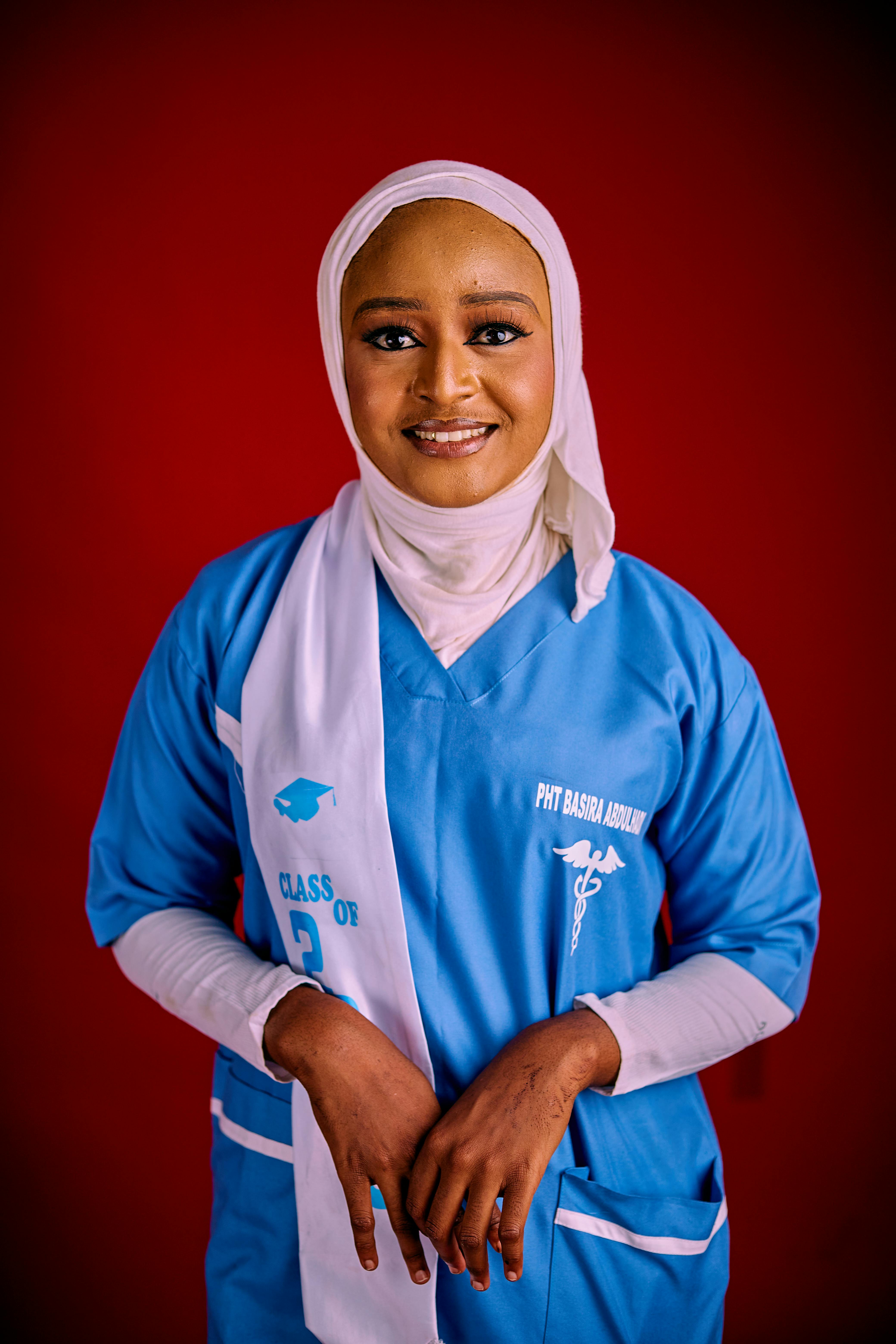 Young female graduate in blue attire and hijab against red backdrop.