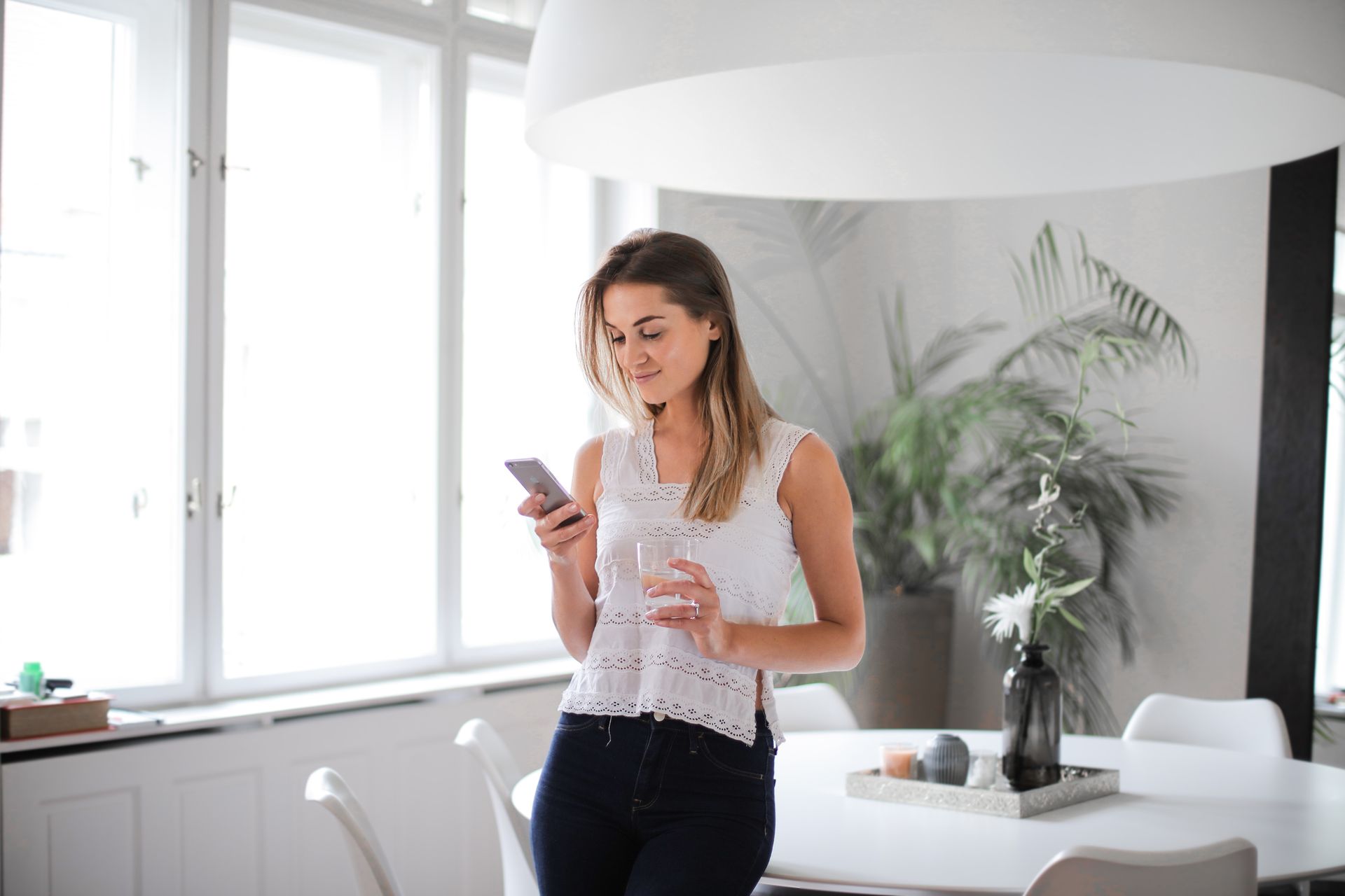 A woman in a white top texting on a smartphone indoors, holding a glass of water.