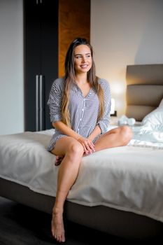 A woman in a striped dress sits happily on a bed in a modern bedroom, smiling warmly.