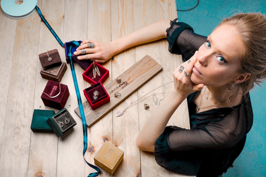 Free From above serious businesswoman looking up at camera while sitting at wooden table with small jewelry boxes with bijouterie and decorating with ribbon as present Stock Photo