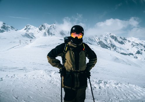 A skier stands confidently in full gear against a backdrop of snow-covered alpine peaks.
